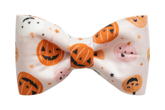 A festive dog bow tie with a pale pink background, decorated with a repeating pattern of cheerful jack-o’-lantern faces. The pumpkins have a variety of playful and spooky expressions, surrounded by small stars for a whimsical Halloween touch.