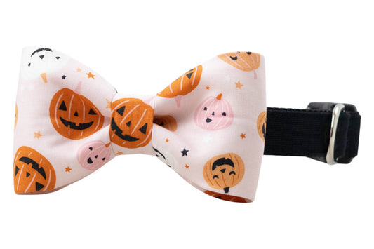 A festive dog bow tie with an adjustable strap and a pastel pink fabric bow tie covered in playful jack-o’-lanterns. The pumpkins appear with a unique carved face, surrounded by scattered stars for a fun Halloween look.