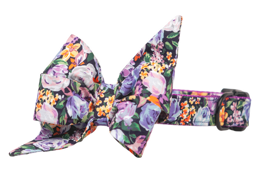 Floral dog collar with an oversized decorative belle bow in matching fabric, printed with multicolored flowers on a dark base. The belle bow has dramatic, pointed ends and a voluminous shape, paired with a D-ring and adjustable fit.