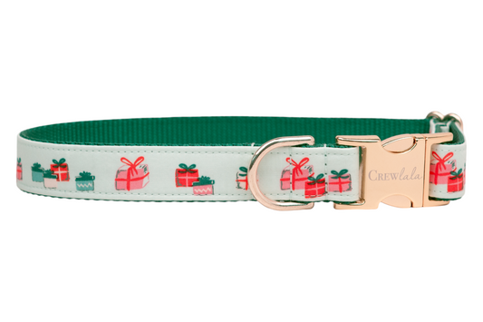 A festive dog collar with a soft green fabric with a repeating pattern of colorful gift boxes each tied with bows. The collar features a sturdy dark green nylon interior, a side-release buckle engraved with the brand name "Crewlala," and a matching D-ring.