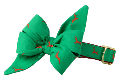 A holiday dog collar with a structured green bow, featuring a repeating reindeer print. The belle bow includes tapered tails and sits atop a matching collar with a nylon backing and metal hardware.