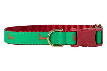 A festive dog collar with a green fabric overlay printed with reindeer silhouettes. The collar is backed with woven webbing and features luxe hardware, including a plastic buckle engraved with “Crewlala.”