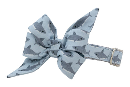Pale blue pet collar with a tied bow detail, decorated with small dark gray sharks and finished with a metal D-ring.