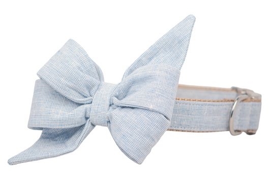 Dog collar in soft light blue linen fabric with a subtle grid pattern, featuring a large matching decorative belle bow. The belle bow is feminine and structured with layered folds and pointed ends, and the collar is finished with silver-tone hardware.