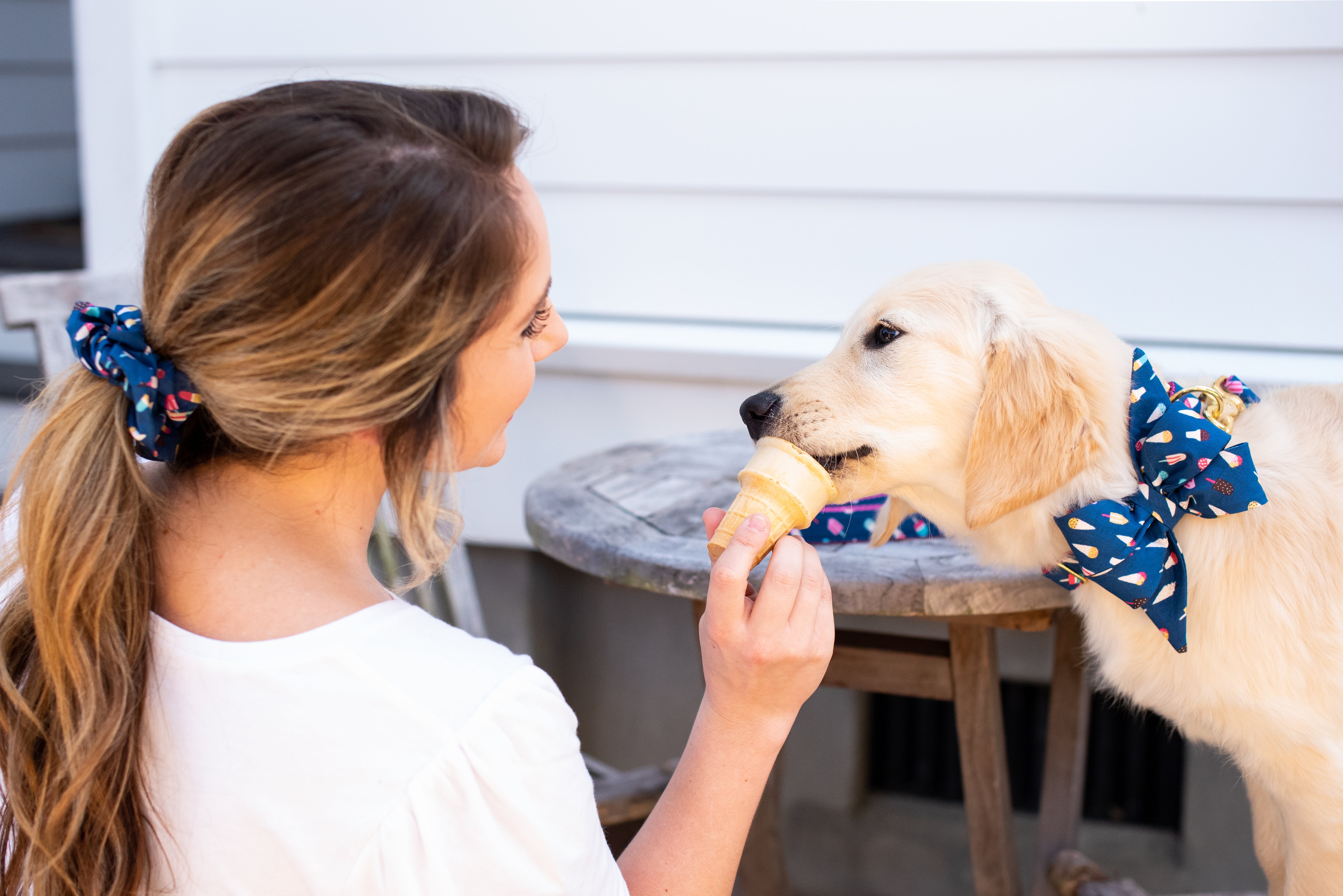 A young woman and a golden retriever puppy share a tender moment as she holds an ice cream cone for the pup to lick. Both wear matching accessories in a navy blue fabric patterned with colorful ice creams and popsicles—the woman has a scrunchie in her ponytail, and the dog wears a large decorative belle bow collar.