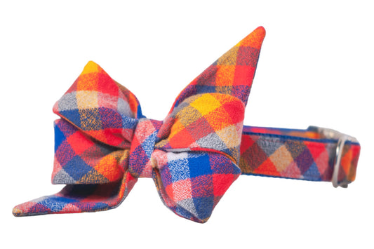 Dog collar with a matching oversized belle bow in a colorful plaid pattern featuring red, orange, blue. The belle bow has pointed tails and crisp folds, while the collar includes metal hardware for adjustment and attachment.