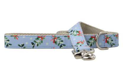 Winter-themed dog leash made from soft blue fabric decorated with mistletoe clusters tied with bows and surrounded by snow-like dots. The underside features nylon webbing for strength, and the leash includes hardware: a rotating clasp and D-ring for attaching accessories.