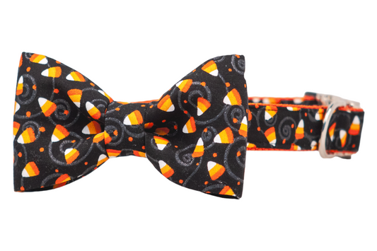 Dog collar with an attached bow tie featuring a festive candy corn pattern. The black fabric is decorated with bright candy corn pieces, whimsical swirls, and polka dots, making it a playful Halloween accessory.