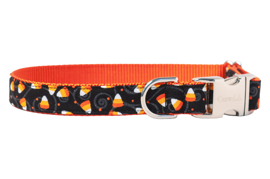 Halloween-themed dog collar with a black fabric base patterned in candy corn pieces and swirling accents. The bright candy corns pop against the dark background.  A metal buckle and D-ring complete the festive design.