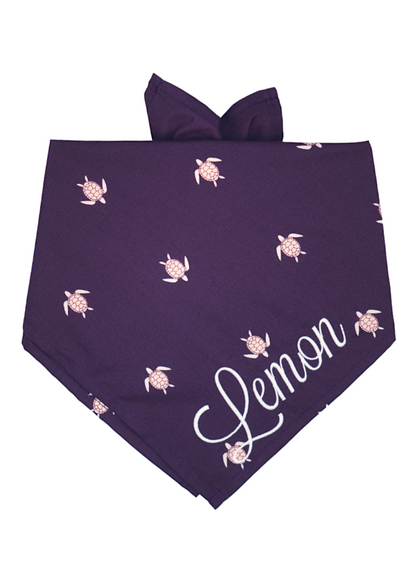 A deep purple dog bandana with a repeating pattern of sea turtles, each with a hexagon-patterned shell and flippers. The bandana is tied at the top and features the name “Lemon” embroidered in large, flowing white script across the bottom corner.