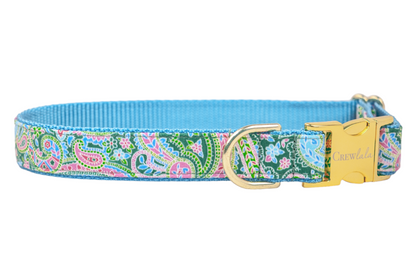 A colorful dog collar featuring a paisley pattern in shades of green, pink, and blue with small floral accents. The collar has a bright blue woven inner lining, a shiny gold D-ring for leash attachment, and a gold side-release buckle engraved with the brand name "Crew Lala."