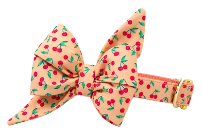 Dog collar featuring a large belle bow in Very Cherry fabric, a peach fabric with small red cherries and green leaves, attached to a matching Very Cherry collar with a nylon inner lining and gleaming gold hardware, including a D-ring and buckle.