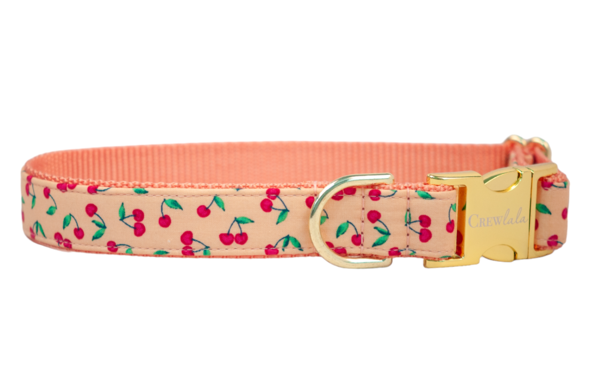 A peach-colored dog collar adorned with a playful cherry print, featuring small red cherries with green stems and leaves. The collar includes a gold metal D-ring and a polished side-release buckle engraved with the brand name "Crew LaLa."