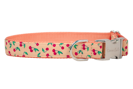 A peach-colored dog collar adorned with a playful cherry print, featuring small red cherries with green stems and leaves. The collar includes a silver metal D-ring and a polished side-release buckle engraved with the brand name "Crew LaLa."