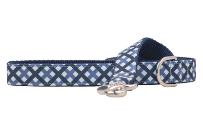 A close-up of a fabric dog leash with a navy blue and light blue plaid pattern on the outside and a textured dark blue inner webbing lining. The leash features reinforced stitching, a shiny metal D-ring, and a swivel clasp for attaching a leash.