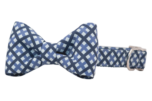 A blue plaid dog bow tie collar with crisp lines and a grid-like pattern of navy and pale blue. The adjustable collar is made of matching fabric and includes a metallic D-ring for function and style.