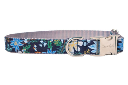 A winter-themed dog collar with a navy blue background, featuring a festive floral pattern of blue poinsettias, light colored flowers, holly leaves, and metallic detailing. The collar includes a metal D-ring and a sturdy metal side-release buckle engraved with the brand name "Crew LaLa." The interior is lined with soft nylon webbing.