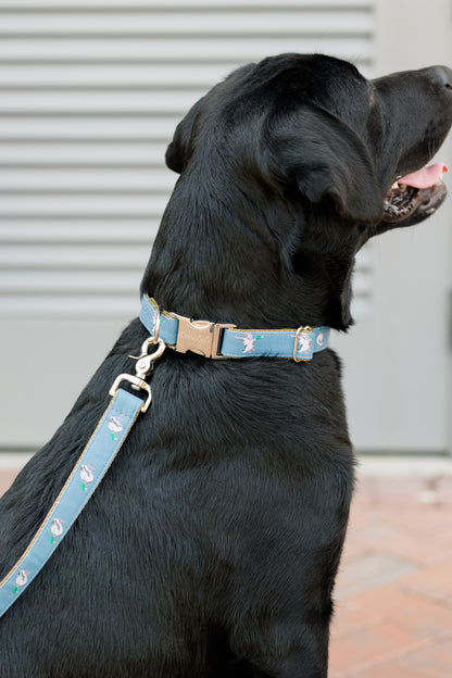 A black Labrador facing sideways, wearing a matching blue collar and leash set decorated with mallard duck illustrations. The collar features a gold-tone buckle engraved with the word “CrewLaLa,” and the leash is clipped to a metal ring at the back of the collar.