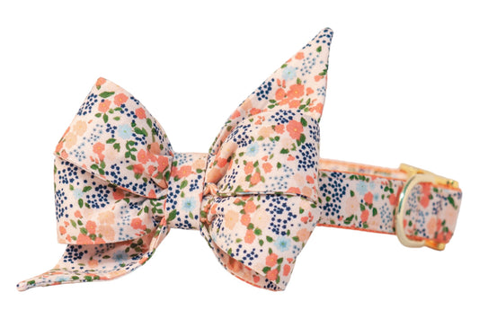 Elegant dog collar featuring an oversized belle bow with pointed ends, patterned with small blossoms, buds, and flowers on a peach background, attached to a matching collar with metal hardware. 