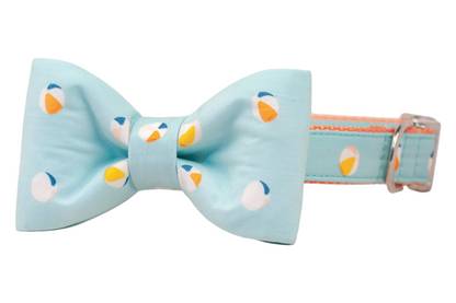A light blue dog collar with an oversized fabric dog bowtie, both printed with a repeating beach ball pattern in orange, yellow, peach, and navy. The bow has wide, full loops and long pointed tails, while the collar features silver hardware.