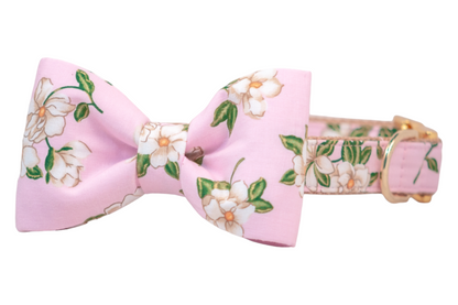 Dog collar crafted from light pink fabric featuring a print of white magnolia flowers with leaves. Attached at the front is a symmetrical bow tie made from matching floral fabric. The collar is backed with durable webbing and finished with a gold-tone buckle and D-ring for leash or tag attachment.