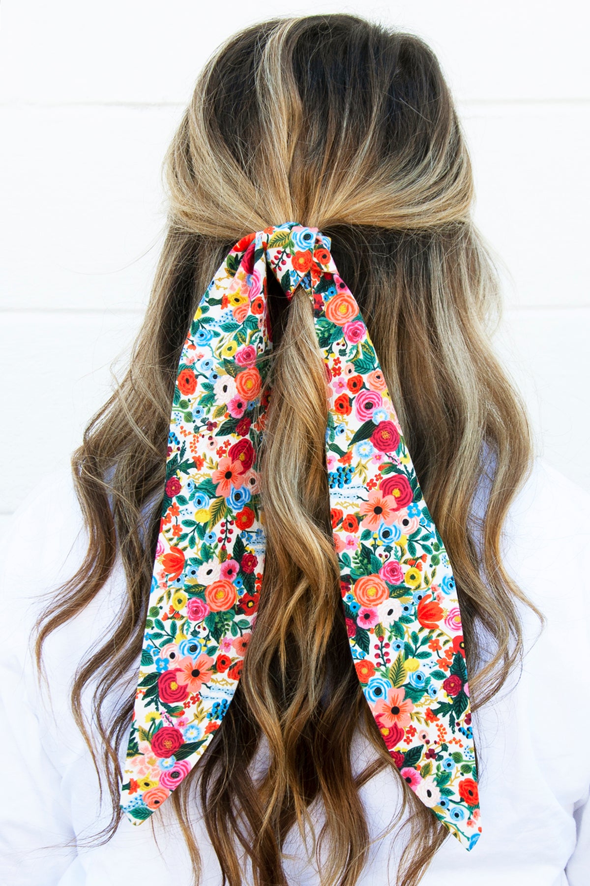 A woman with long, wavy, dark blonde hair wears a vibrant floral hair bow tied at the back of her head. The fabric features a colorful mix of red, pink, blue, yellow, and green flowers, with two long tails draping down over her hair.