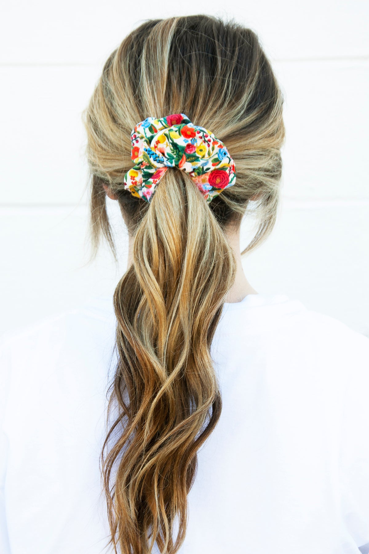 The back view of a person with loosely curled, light brown hair secured in a ponytail using a fabric scrunchie covered in a multicolored floral print. The scrunchie has a voluminous, ruffled appearance.