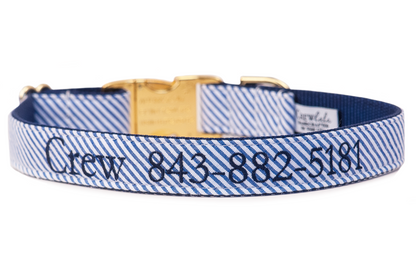A customized blue and white striped seersucker dog collar with dark navy diagonal stripes and navy blue inner webbing. The collar features navy embroidered text reading "Crew 843-882-5181" across the front. It includes a gold-tone side-release buckle and matching D-ring, adding a polished, upscale touch. The design combines practical identification with preppy, nautical style.