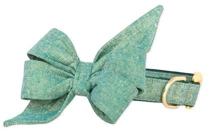 A green fabric dog collar featuring a large, structured belle bow with pointed tails, made from textured, woven material with specks of glitter. The matching collar is equipped with a metal D-ring and buckle, adding a refined touch to the stylish accessory.