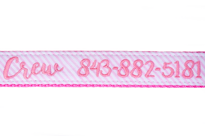 Close-up of a pink and white seersucker dog collar with bright pink embroidered text reading “Crew 843-882-5181.” The stitching is neatly sewn into the fabric, framed by hot pink webbing and double-stitched edges.