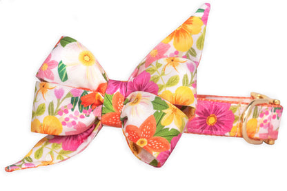 Dog collar with a matching oversized feminine belle bow in a tropical floral pattern, featuring bright orange lilies, pink cosmos, yellow daisies, and green leaves on a soft white background. The collar includes gold-tone hardware, a D-ring for leash attachment, and matching floral print throughout.