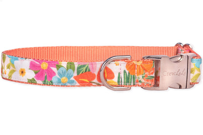 A colorful dog collar made from fabric printed with vibrant summer flowers—such as orange lilies, fuchsia daisies, yellow blooms, and pink clusters—set against a pale background. It includes a silver-tone buckle clasp engraved with “Crew LaLa,” a matching D-ring.