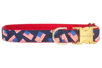 Adjustable dog collar with a navy blue fabric overlay featuring a repeating pattern of American flags. The collar is backed with vibrant red nylon and includes a polished gold metal buckle engraved with the brand name "Crew LaLa" alongside a matching gold D-ring for attaching tags or a leash.