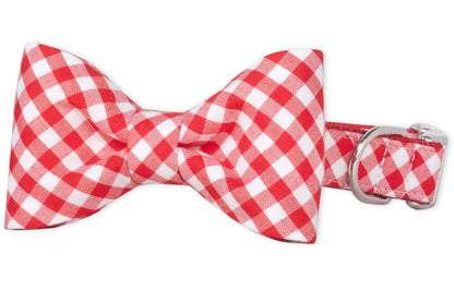 A dog collar with an attached symmetrical bow tie in red plaid fabric. The bow tie is evenly pleated with softly rounded edges, and the collar features a metal D-ring and side-release buckle for secure fastening.