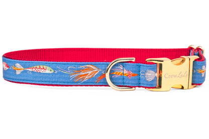 A close-up of a blue dog collar with a red interior featuring a playful fishing lure print, including hooks, feathers, and striped bait. The collar is finished with a shiny gold side-release buckle engraved with "CrewLala" and a sturdy D-ring for leash attachment.