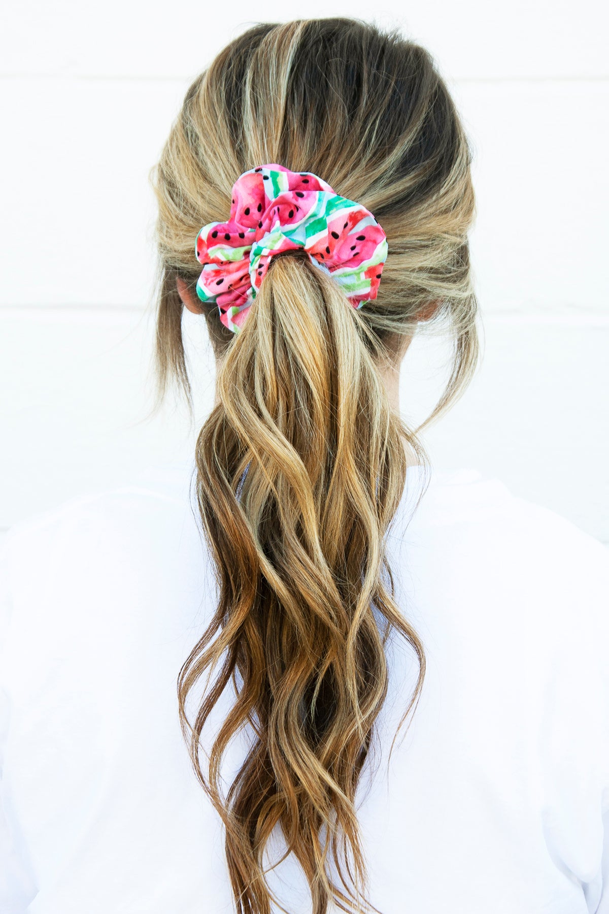 A woman with long, wavy, blonde-highlighted brown hair is wearing a scrunchie in a ponytail. The scrunchie features a vibrant watermelon pattern with pink-red fruit, green-striped rind, and black seeds on a light blue background, adding a playful, summery touch to her hairstyle.